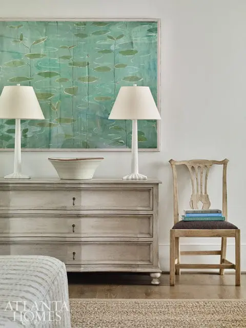 Light gray dresser with three drawers topped with two white lamps and a large framed artwork of a pond with lily pads. A wooden chair with books stacked on the seat sits to the right.