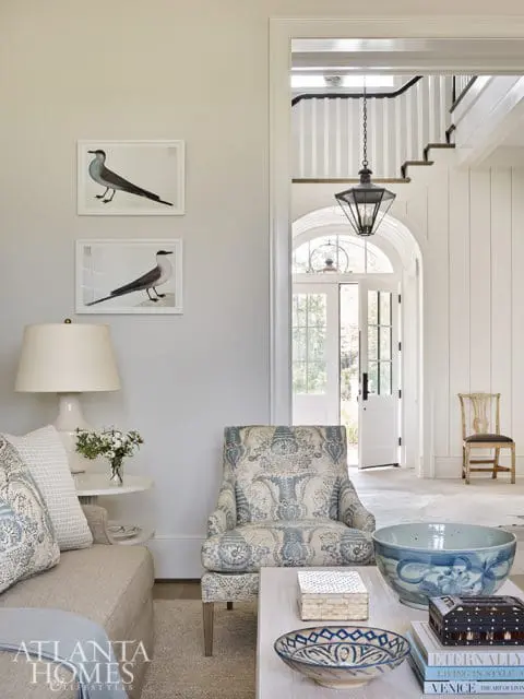 Living room with a neutral sofa, armchair upholstered in blue and white floral fabric, and a coffee table with decorative bowls and books. Two framed bird prints hang on the wall.