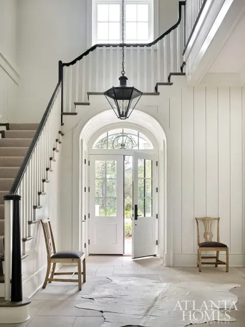 Foyer with a grand staircase, black railing, carpeted stairs, and a large black lantern chandelier. Double doors open to the outside, with two wooden chairs and a cowhide rug on the stone floor.