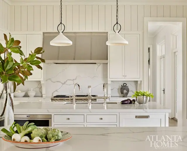 White kitchen with paneled walls, marble countertops and backsplash, stainless steel range hood, and two white pendant lights over the island.