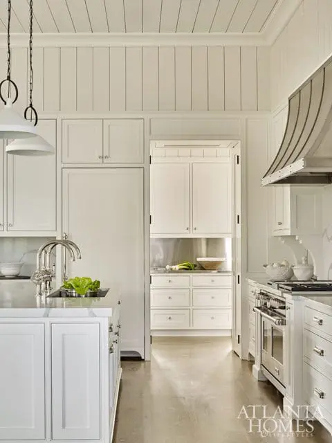 White kitchen with paneled walls and ceiling, marble island with sink, stainless steel range with curved hood, and view into pantry with white cabinetry and stainless steel backsplash.