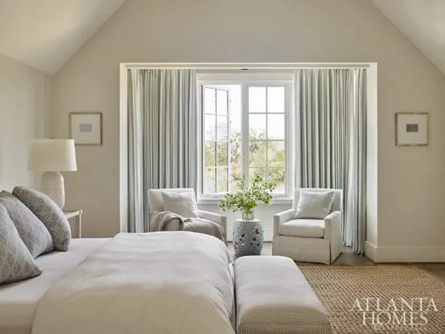 Bedroom with vaulted ceiling, neutral walls, and large window with striped curtains. Two armchairs flank the window, with a small table between them. A bed with white linens is in the foreground.