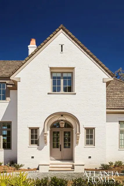 White brick house with arched entryway, double doors, and multiple windows under a brown shingle roof.