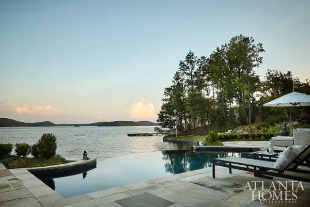 Infinity pool overlooking a lake at sunset, with lounge chairs and a white umbrella on a stone patio. Tall trees line the shore.