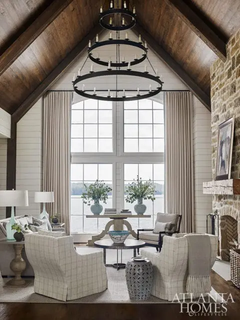 Living room with vaulted wood-paneled ceiling, large arched windows overlooking a lake, stone fireplace, and plaid upholstered chairs.