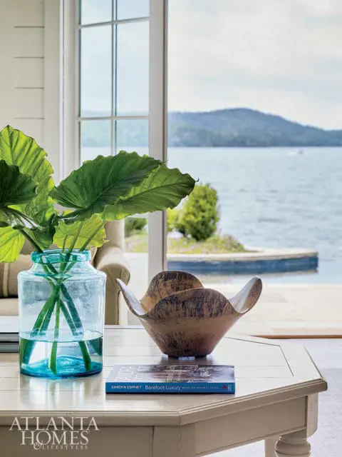 Light wood coffee table with a blue glass vase of large green leaves, a wooden bowl, and a book titled 'Barefoot Luxury Mexican Resort Living'