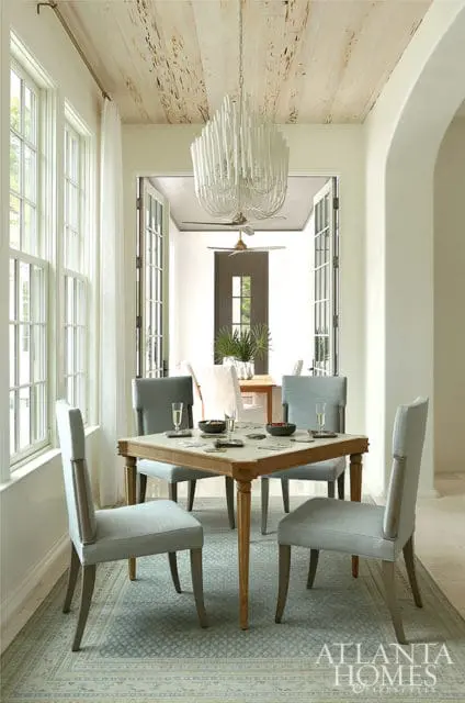 Dining room with a square game table surrounded by four upholstered chairs, a patterned rug, and a white branched chandelier hanging from a distressed wood plank ceiling.