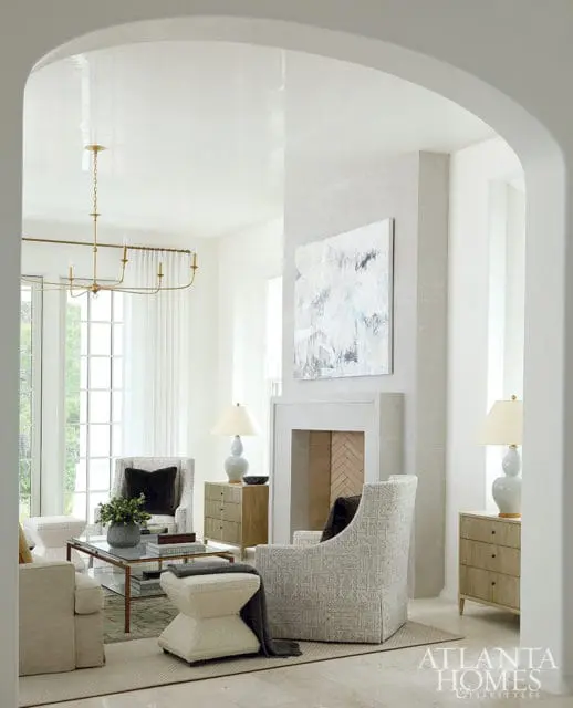 Living room with a light gray fireplace, abstract art, patterned armchairs, a glass-top coffee table, and two wooden nightstands with lamps.