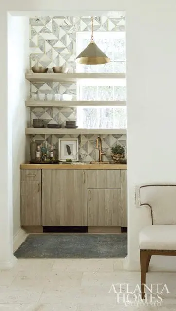 Wet bar with geometric marble backsplash, wood cabinetry, open shelving, brass faucet, and pendant light. A chair is visible to the right.