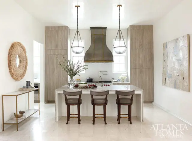 Modern kitchen with light wood cabinetry, a large metal range hood, and a marble island with three leather stools. A large abstract painting hangs on the wall.