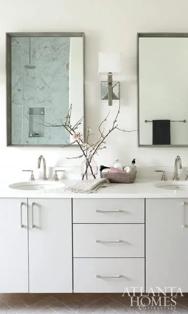 White double vanity with silver hardware, two sinks, and marble shower visible in mirror. A glass vase with branches and pink blossoms sits on the counter.