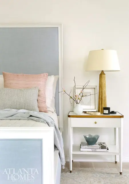 Bedroom with a light blue upholstered headboard, pink and gray throw pillows, and a white nightstand with a gold fluted lamp and decorative objects.