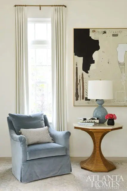 Light blue velvet armchair with throw pillows next to a hexagonal side table with books, a blue ceramic lamp, and a vase of pink roses.
