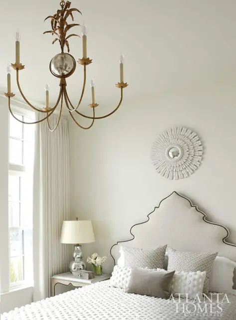 Bedroom with a cream upholstered headboard outlined in black trim, white bedding, and a gold chandelier. A sunburst mirror hangs on the wall above the headboard.