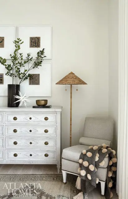 White dresser with brass hardware, framed art, and a floor lamp with a woven shade next to a gray armchair with a pom-pom throw