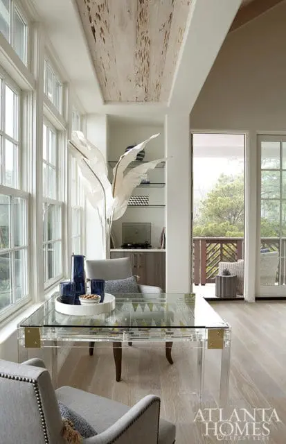 Home office with a clear acrylic desk, gray upholstered chairs, and a wood-paneled ceiling. Large windows overlook a balcony with trees.