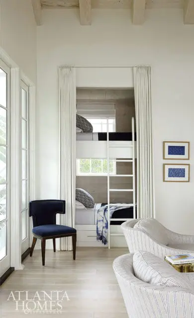 Bunk beds with white ladders and navy bedding in a room with light wood floors, a navy accent chair, and striped chaise lounges.