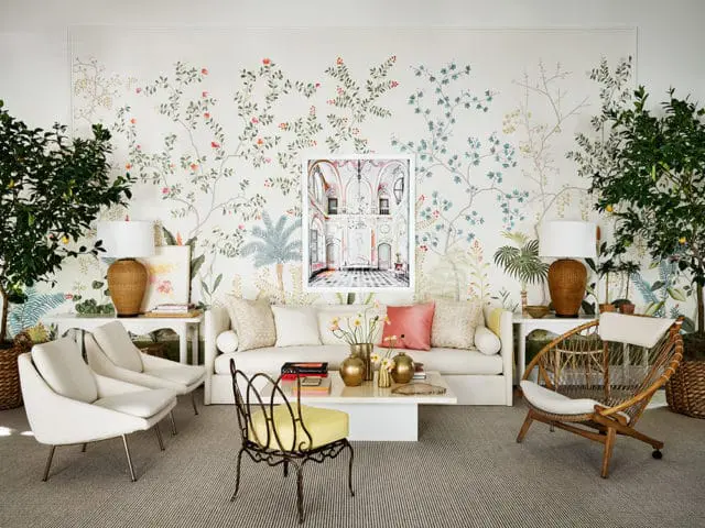 Living room with a large floral and botanical wallpaper, a white sofa, two white armchairs, a rattan accent chair, and a coffee table with books and vases.