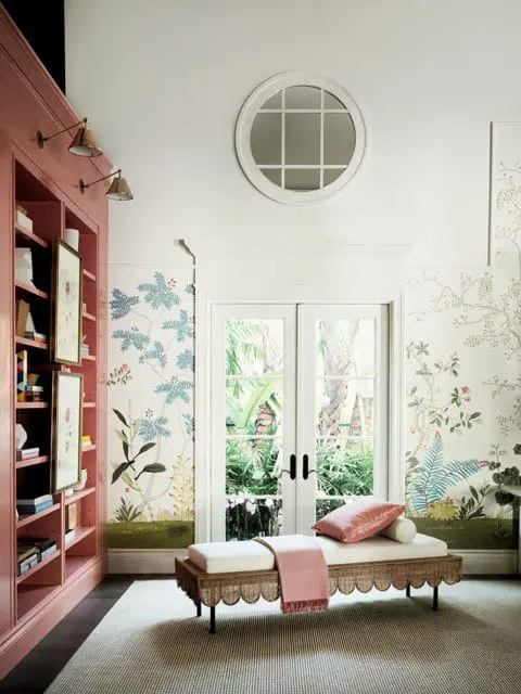 Interior room with a pink built-in bookshelf, botanical wallpaper, a round window, and a wicker daybed with pink pillows and a throw.