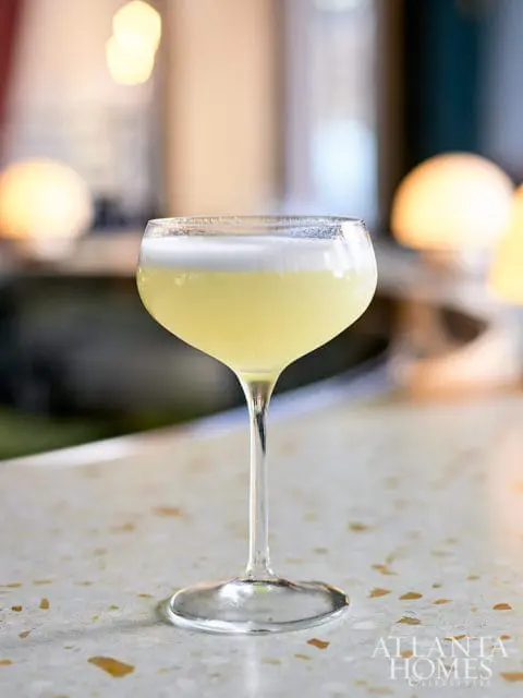 Close-up of a pale yellow cocktail with white foam in a coupe glass, resting on a speckled terrazzo countertop.