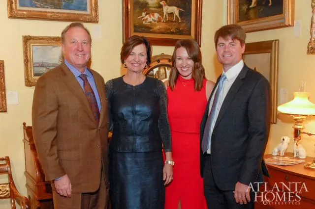 Four people pose for a photo in front of framed artwork and antique furniture. A man in a brown suit stands to the left, followed by a woman in a black leather dress, a woman in a red dress, and a man in a dark suit.
