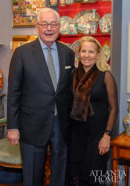 Man in a dark suit and patterned tie stands next to a woman in a black dress and fur stole. Behind them are shelves displaying decorative plates and artwork.