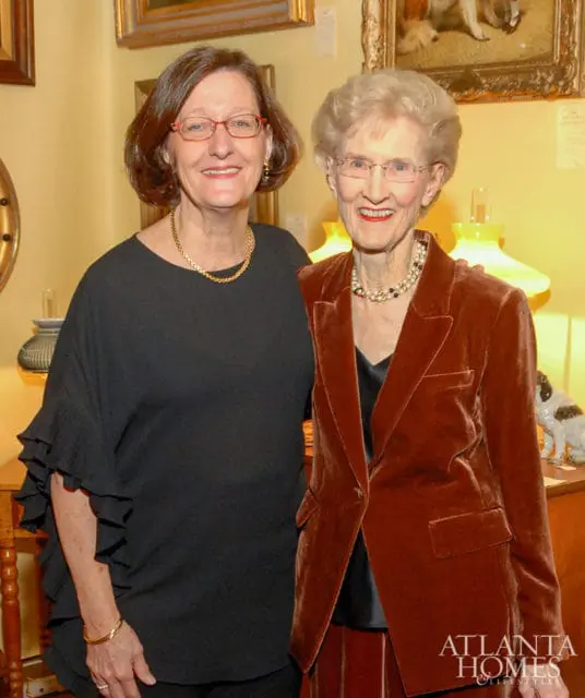 Two women smiling, one in a black ruffled top and gold necklace, the other in a rust-colored velvet blazer and pearl necklace.
