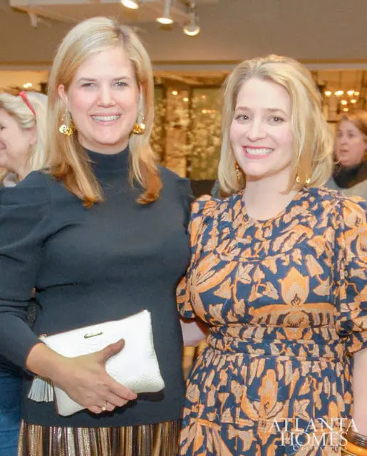 Two women smiling, one in a black turtleneck and metallic skirt holding a white clutch, the other in a blue and orange patterned dress.