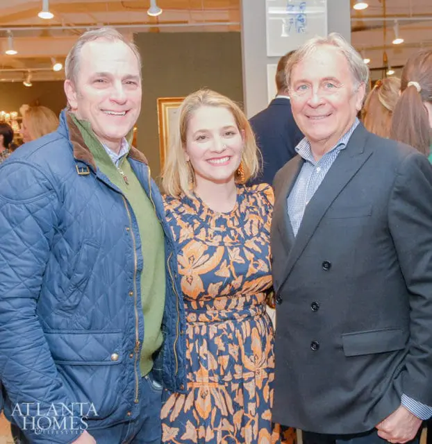 Three people smiling for a photo, including a man in a blue quilted jacket and green sweater, a woman in a navy and orange patterned dress, and a man in a black suit jacket.