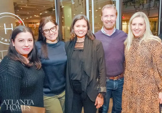 Five people pose for a photo, including two women in the foreground wearing a black sweater and a navy blue top, and three people in the background wearing a black jacket, a purple sweater, and a leopard print dress.