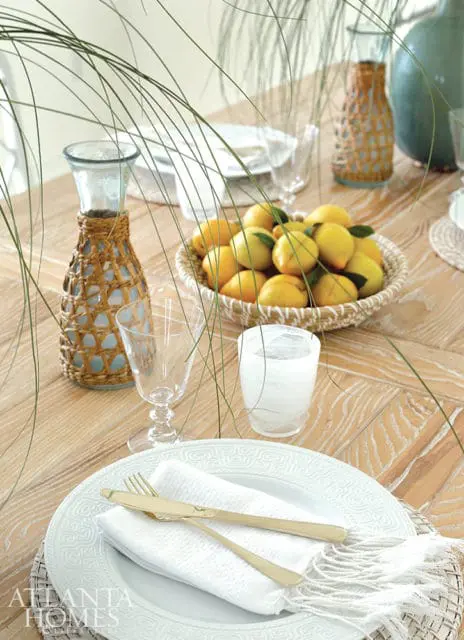 Dining table set with white plates, gold cutlery, a basket of lemons, and a woven-covered glass carafe.