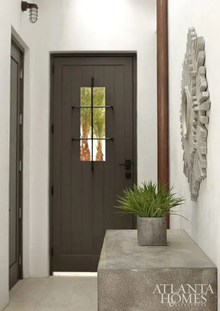 Dark gray paneled door with a small window overlooking palm trees, flanked by white walls and a concrete console table with a potted plant and sunburst mirror.