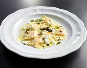 Plate of cheese ravioli topped with sauce, crumbled cheese, herbs, and a drizzle of dark sauce on a white scalloped plate.