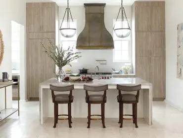 Modern kitchen with light wood cabinetry, a large metal range hood, and a marble island with three upholstered bar stools.