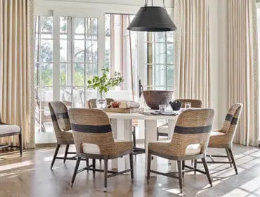 Dining room with a round white table surrounded by upholstered chairs featuring woven natural material and dark horizontal stripes. Large windows with sheer curtains frame the space.