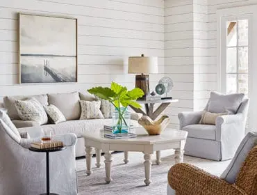 Living room with white shiplap walls, a neutral-toned sofa and armchairs, a pale wood octagonal coffee table, and a framed artwork of a pier over water.