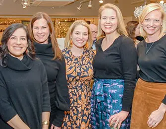 Five women smiling at the camera, dressed in casual attire. The woman second from right wears a black sweater and blue skirt with a purple tiger pattern.