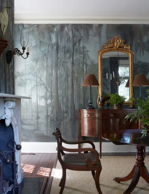 Dining room with a mural wallpaper depicting a swamp scene, a marble fireplace, antique wooden furniture, and a gold ornate mirror.