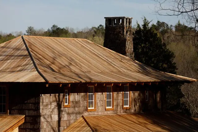 Wooden shingle exterior walls and a sloped wooden plank roof with a stone chimney on a house surrounded by trees.