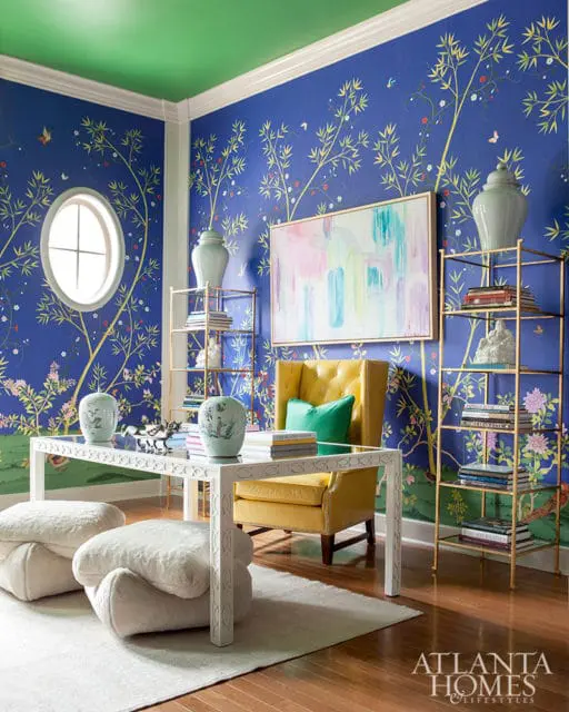 Blue Chinoiserie wallpaper adorns the walls of a room with a green ceiling, featuring a yellow wingback chair, white desk, and gold shelving units.