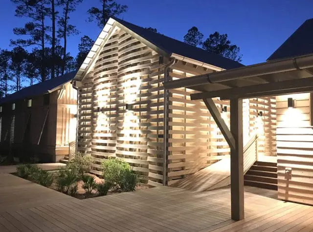 Modern wooden building with slatted facade illuminated at dusk, featuring a ramp and wooden deck in the foreground.
