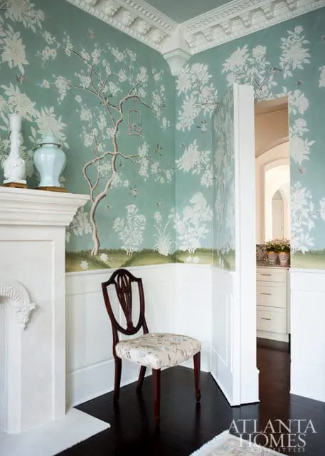 Corner of a room with a fireplace, a wooden chair upholstered in floral fabric, and walls covered in a chinoiserie-style wallpaper featuring trees, flowers, and birds.
