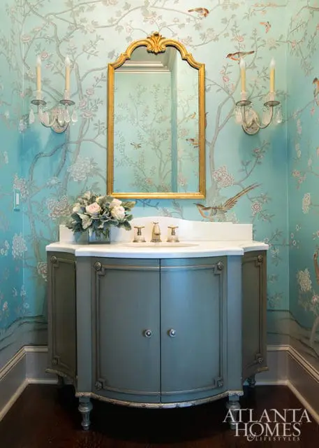 Powder room with blue Chinoiserie wallpaper featuring birds and branches, a gold ornate mirror, and a gray vanity with a white marble countertop.