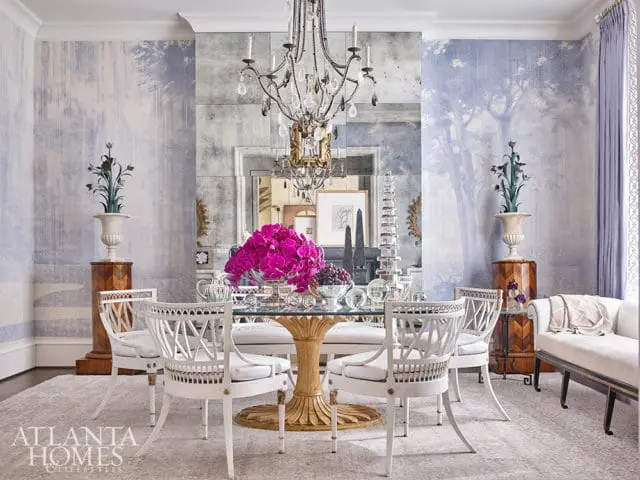 Dining room with blue toile wallpaper, mirrored wall, crystal chandelier, glass-top dining table with white chairs, and a white bench.