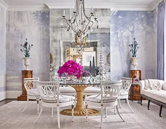 Dining room with a round glass-top table, ornate chandelier, and white upholstered chairs. Walls feature a scenic mural and a large antique mirror.