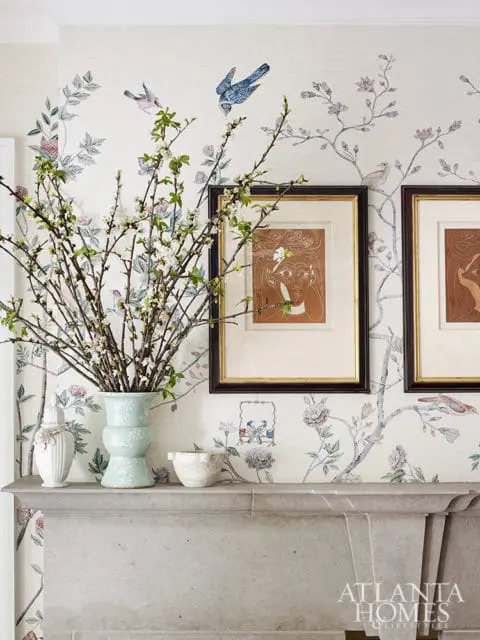 Stone fireplace mantel topped with a pale blue vase of branches, a white urn, and a decorative bowl, set against wallpaper with a bird and floral motif.