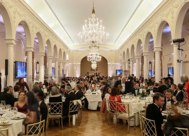 Formal dining hall with guests seated at round tables, ornate columns, arched doorways, and large crystal chandeliers overhead.