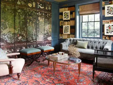 Library with dark blue walls, floor-to-ceiling bookshelves, a large ornate screen depicting cranes and foliage, tufted gray sofa, upholstered armchairs, and a patterned red rug.