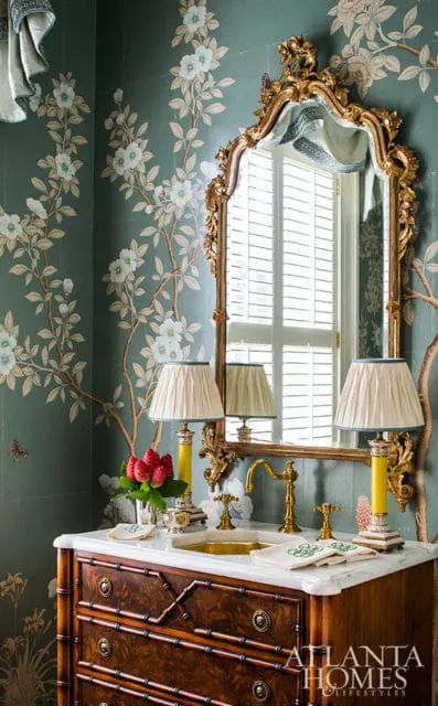 Powder room with teal floral wallpaper, a dark wood bamboo-style vanity, marble countertop, brass fixtures, a gold ornate mirror, and two yellow lamps with pleated shades.