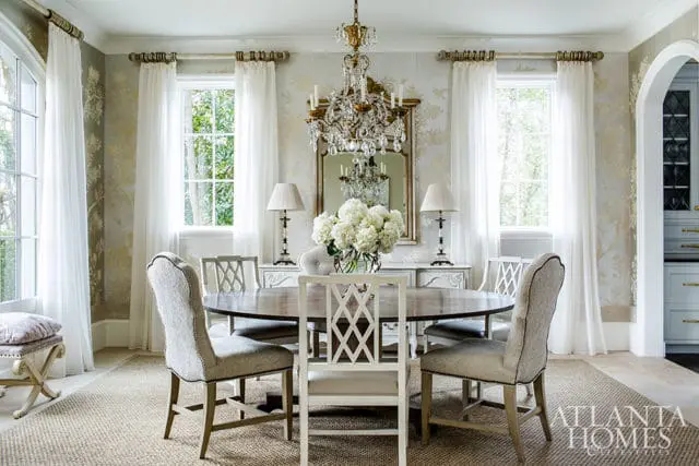 Formal dining room with a round wooden table, upholstered chairs, a crystal chandelier, and a mirrored buffet with lamps and floral arrangement.